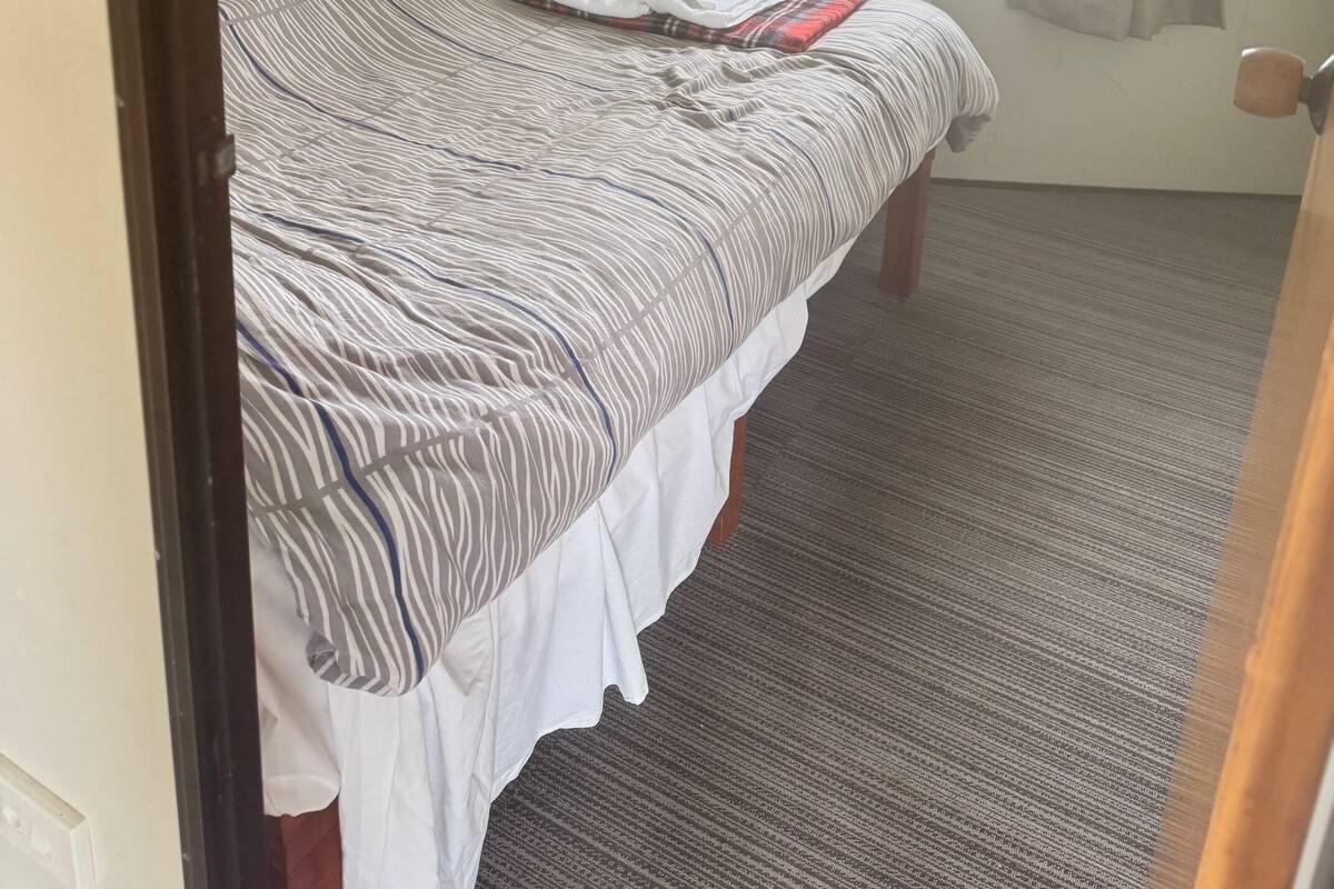 Quarto pequeno com uma cama de solteiro, coberta por um edredom listrado, uma janela com cortinas finas e vista para flores amarelas lá fora.