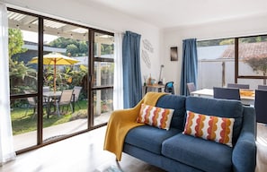 A modern living room with a sofa, a dining table, and a view of a garden through large windows.