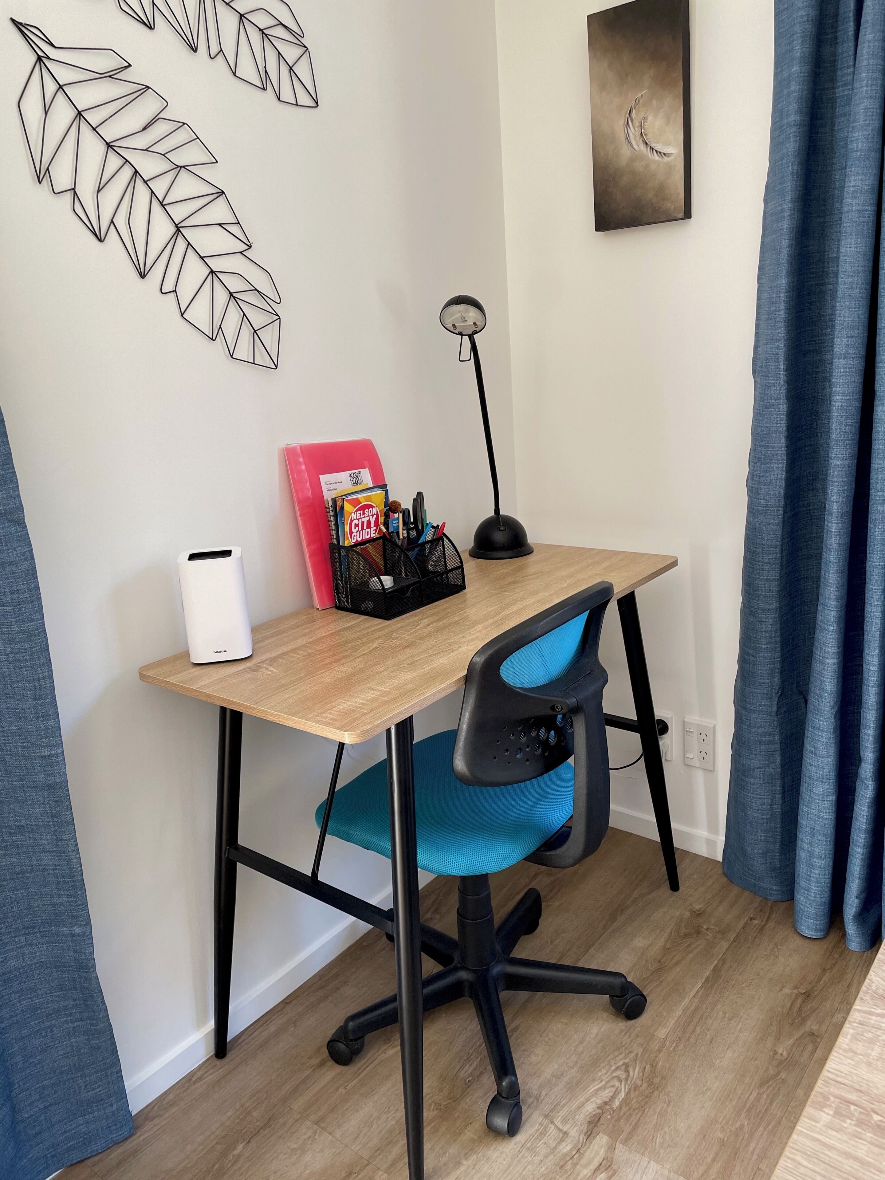 Un espacio de trabajo con un escritorio de madera, una silla de oficina azul, una lámpara blanca y una obra de arte en la pared.