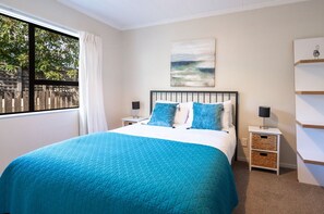 A bedroom with a bed, two bedside tables, a window with curtains, a wall-mounted artwork, and a white shelving unit.