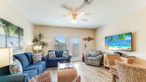 A living room with a blue sofa, wicker chairs, a coffee table, and a television displaying a beach scene. - Family Getaway | Heated Pool, Playground & Beach Close By (Corpus Christi)