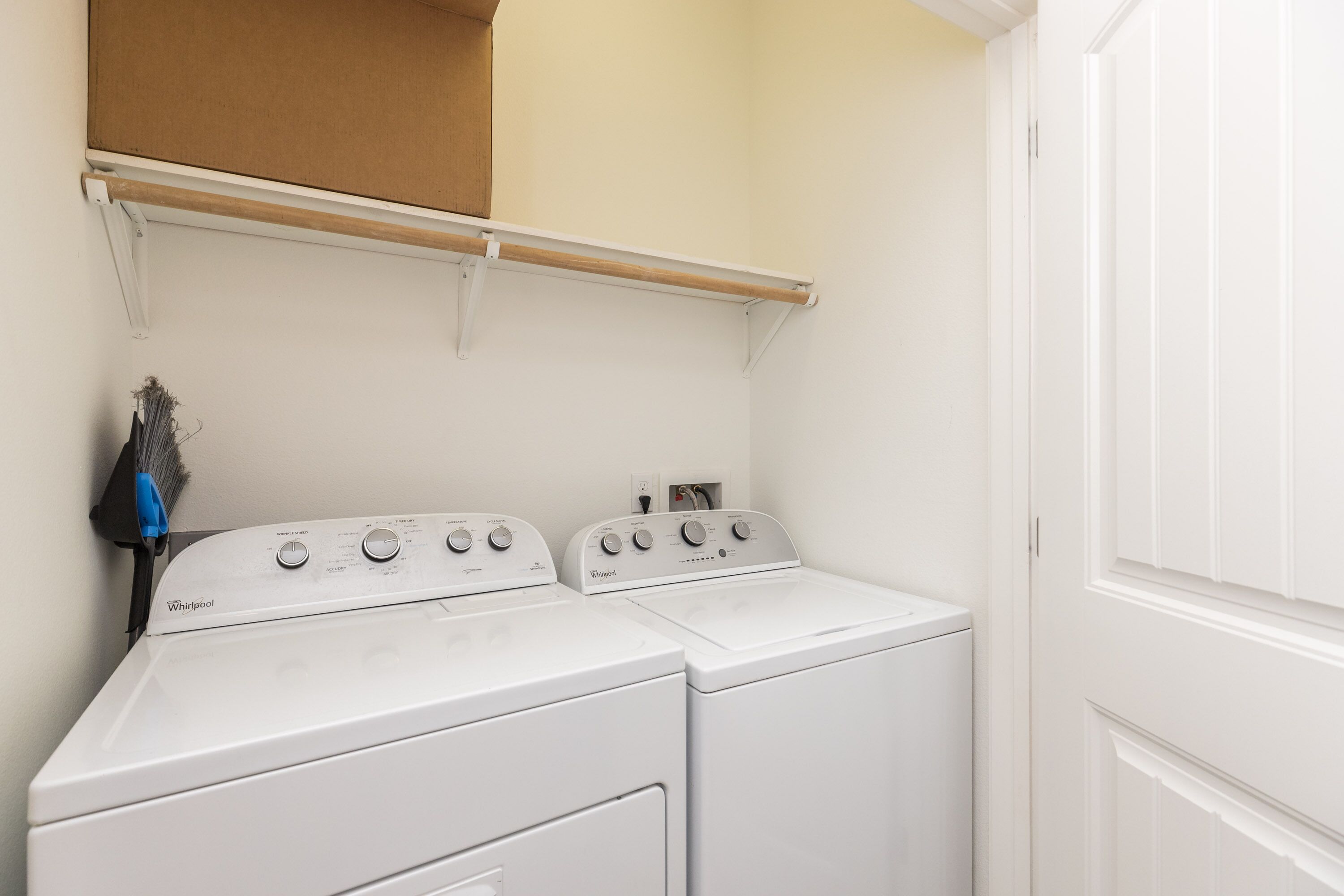 A laundry room with two stacked washer and dryer units, a wooden shelf above, and a broom leaning against the wall.