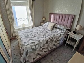 Un dormitorio con cama, mesitas de noche, una ventana con cortinas y cabecero.