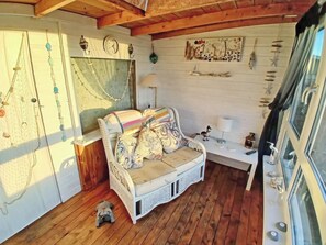 A cozy room with a wooden floor, a white sofa with patterned cushions, a small table with a lamp, and wall decorations including a clock and a framed artwork.