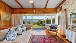 A cozy living room with a large window, wooden ceiling beams, a sofa with patterned cushions, a small table, and a view of the outdoors.