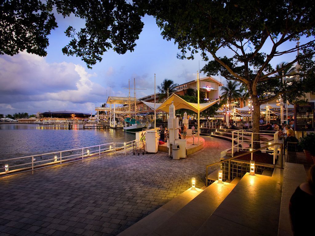 A waterfront promenade with a modern building, a boat dock, and a seating area with tables and chairs.