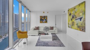 Un salon moderne avec un canapé blanc, un fauteuil jaune et une table basse. Un grand tableau abstrait orne le mur, et de larges fenêtres offrent une vue sur le paysage urbain.