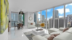 Un salon moderne avec un canapé blanc, un coin repas comprenant une table et des chaises, et de grandes fenêtres offrant une vue sur la ville.
