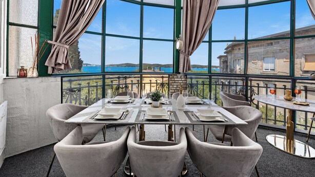 Un'area pranzo con un tavolo apparecchiato per sei persone, ampie finestre con tende e vista sul mare.