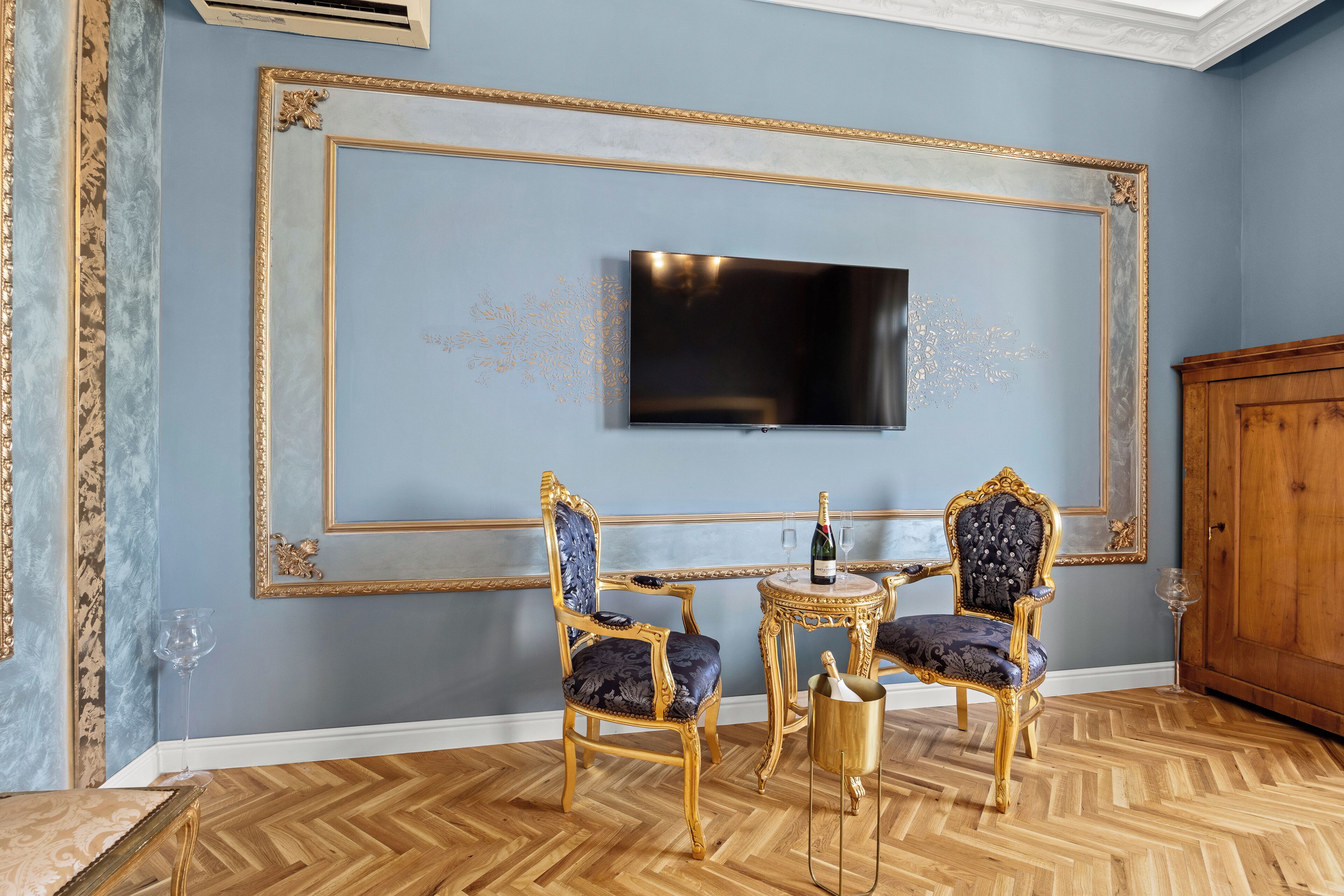 A room with a large flat-screen TV mounted on a blue wall, two ornate chairs with gold frames and blue upholstery, a small round table with a bottle and glass, and a wooden cabinet.