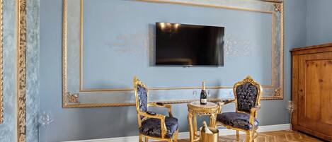 A room with a large flat-screen TV mounted on a blue wall, two ornate chairs with gold frames and blue upholstery, a small round table with a bottle and glass, and a wooden cabinet.