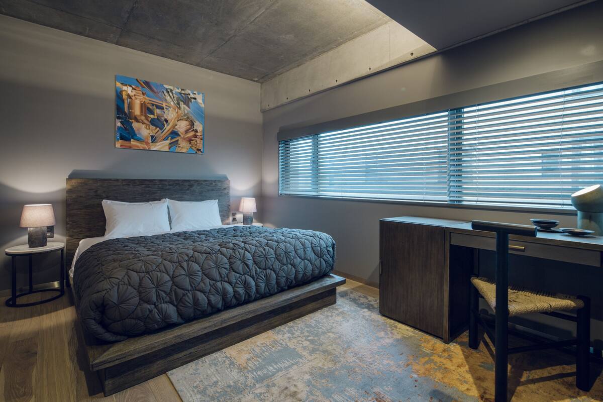 A modern bedroom with a large bed, a desk, and a window with blinds.