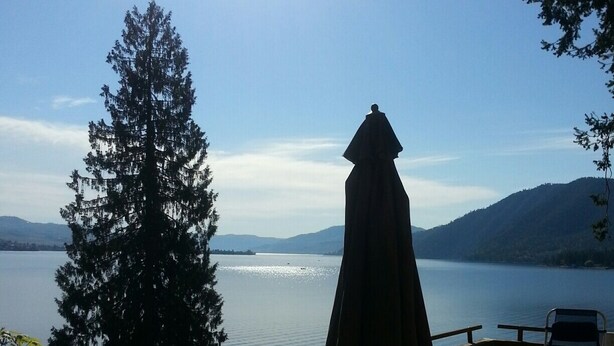 A wooden deck with a table and chairs overlooking a lake, with a large umbrella and a view of distant mountains.