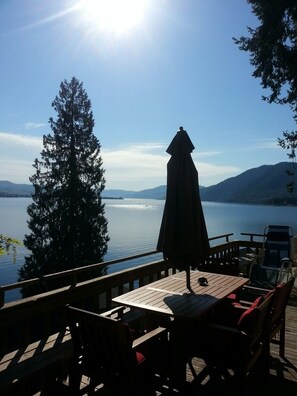 A wooden deck with a table and chairs overlooking a lake, with a large umbrella and a view of distant mountains.