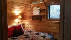 A cozy wooden cabin bedroom with a bed, pillows, a nightstand, a lamp, shelves with towels, and a window with a view of trees.