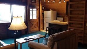 A cozy log cabin interior with a dining area, a kitchen with a refrigerator and stove, a wooden door, and a lamp.
