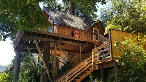 A wooden treehouse with a balcony and stairs, surrounded by lush greenery.