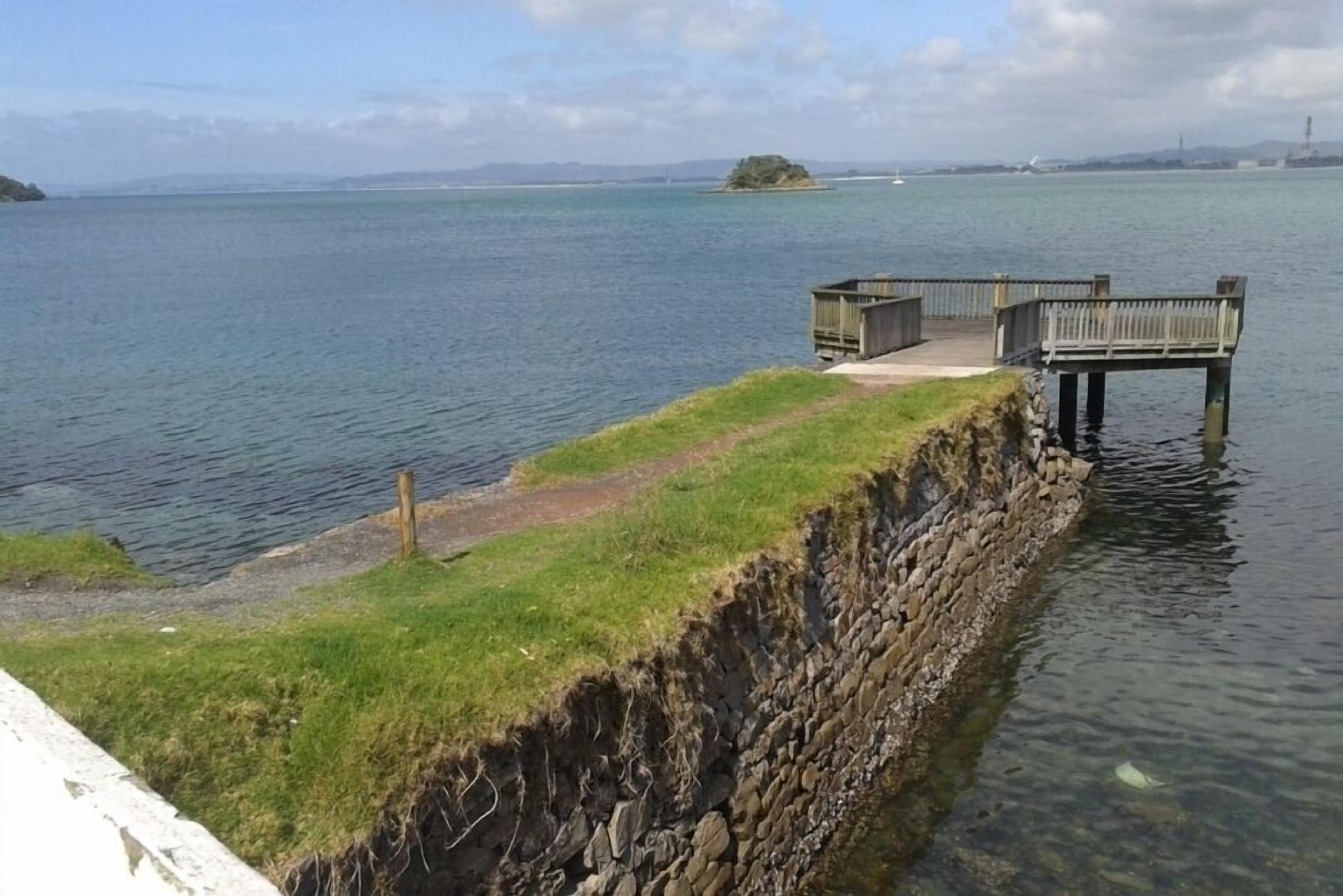 Una propiedad frente al agua con un área verde, un muro de contención de piedra y un muelle de madera que se adentra en el agua.