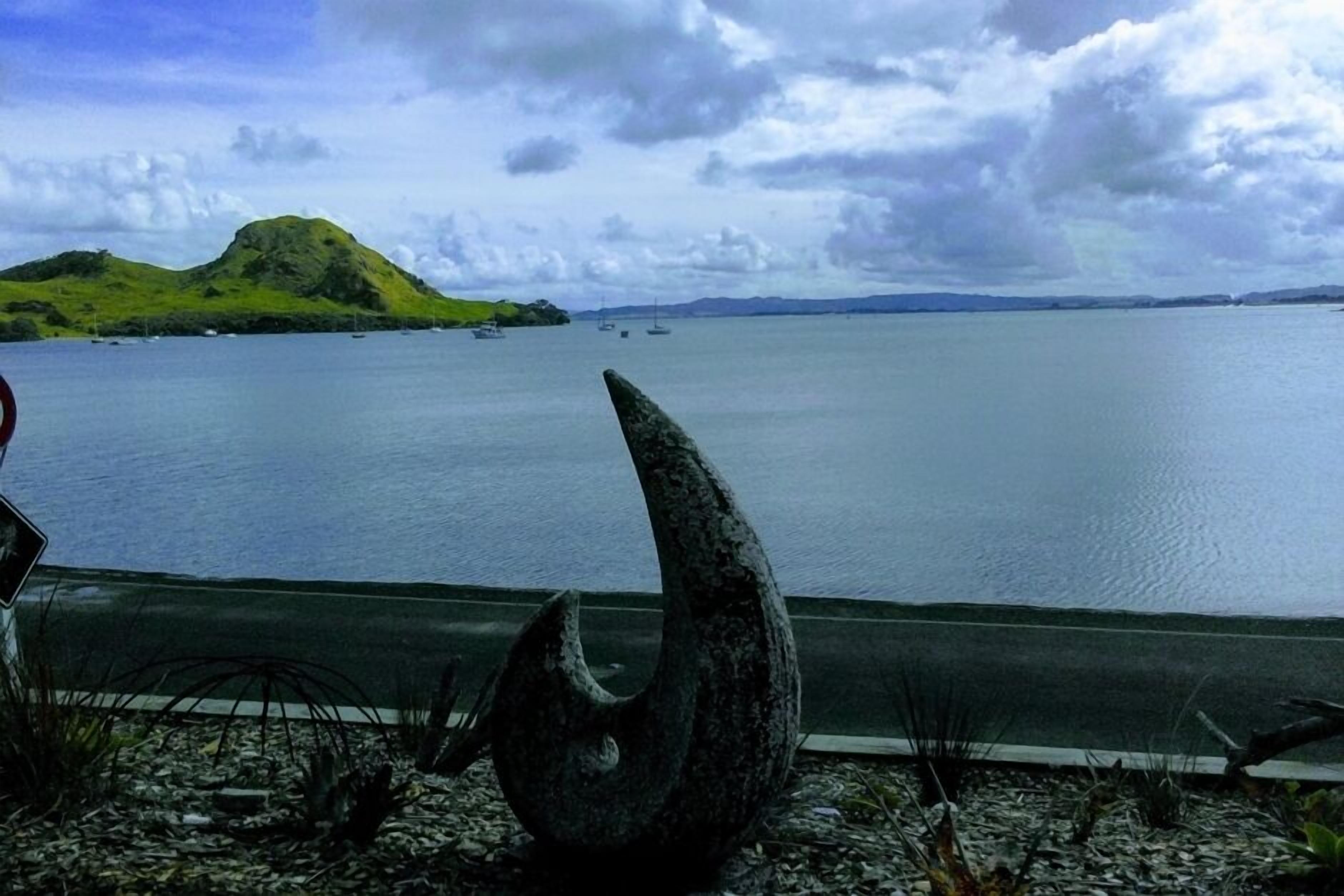 Vista costera con una extensa masa de agua, una colina verde y una escultura en primer plano.