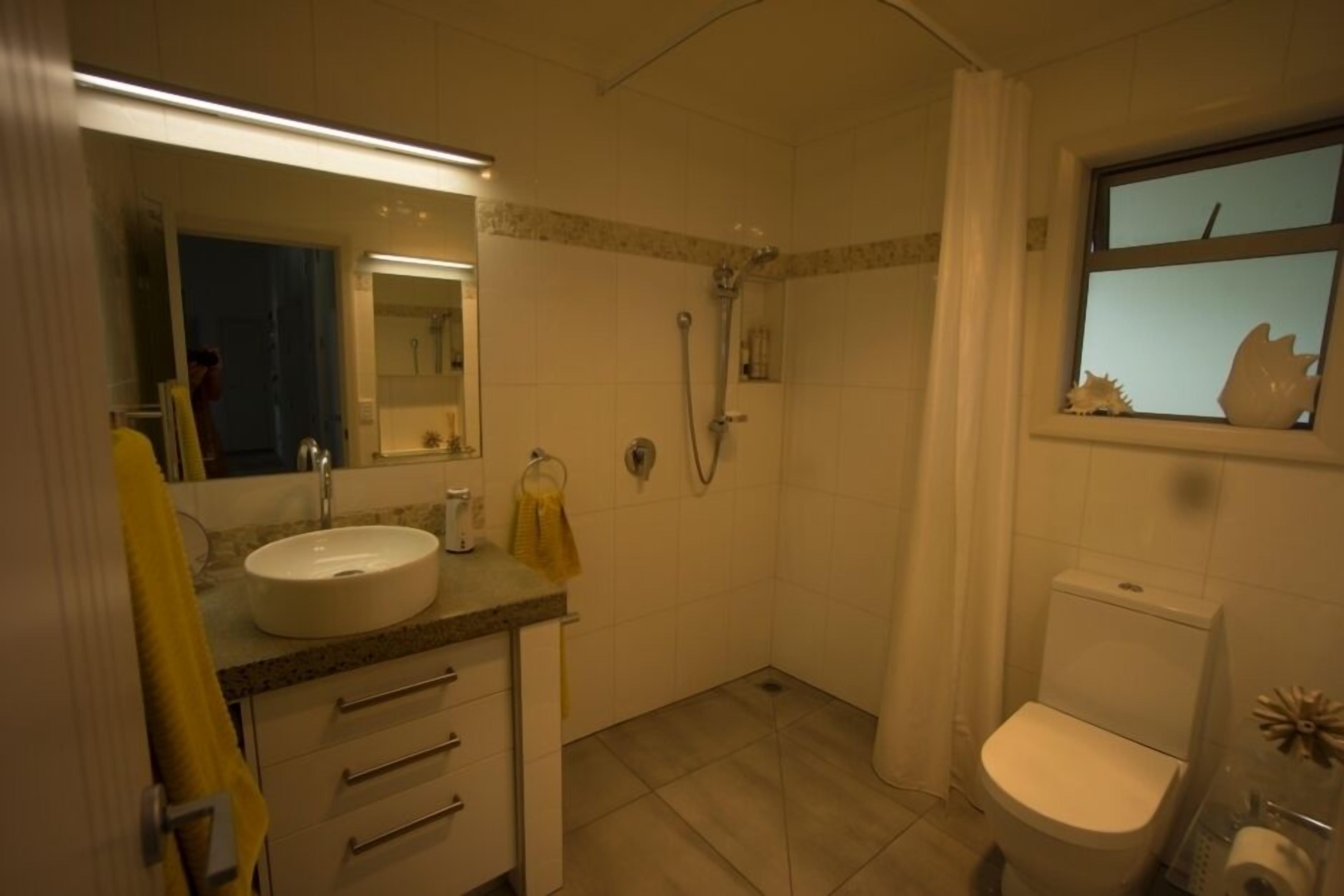 Un bagno con lavandino, specchio, doccia, WC e una finestra con un oggetto decorativo.