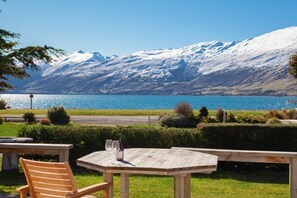 Una mesa de madera con dos sillas y una botella de vino, con vista a un lago y una montaña nevada al fondo.