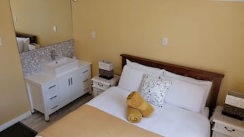 Habitación de hotel ordenada con cama, mesita de noche, espejo y baño con lavabo y tocador.