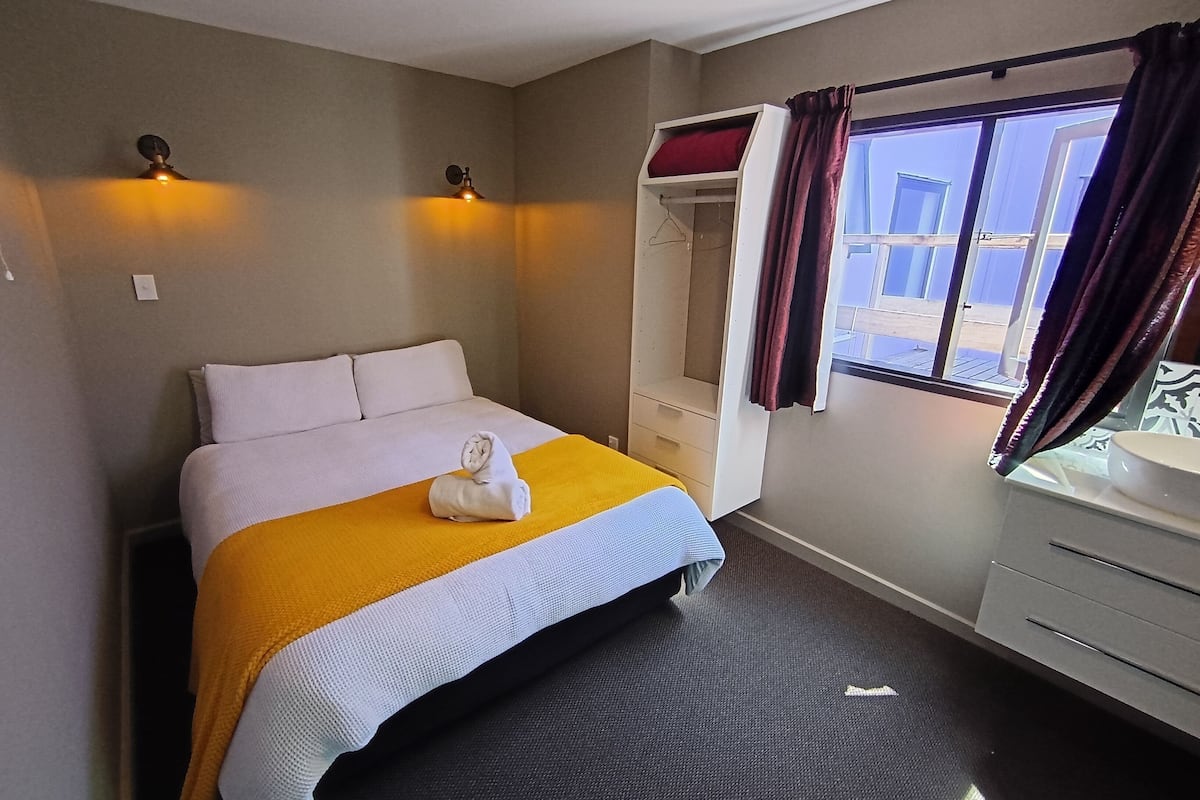 A neatly made bed with a yellow blanket and a white towel, a window with curtains, and a wardrobe with a mirror.