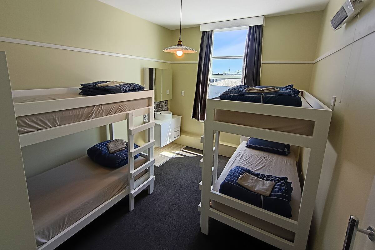 A dormitory room with bunk beds, a small desk, a mirror, and a window with curtains.