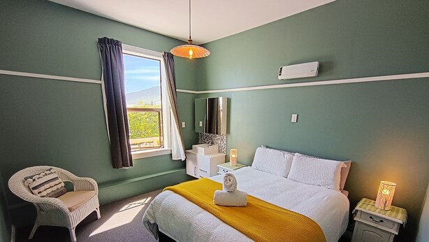 Un dormitorio con cama, una silla, una mesita de noche con lámpara y una ventana con cortinas.