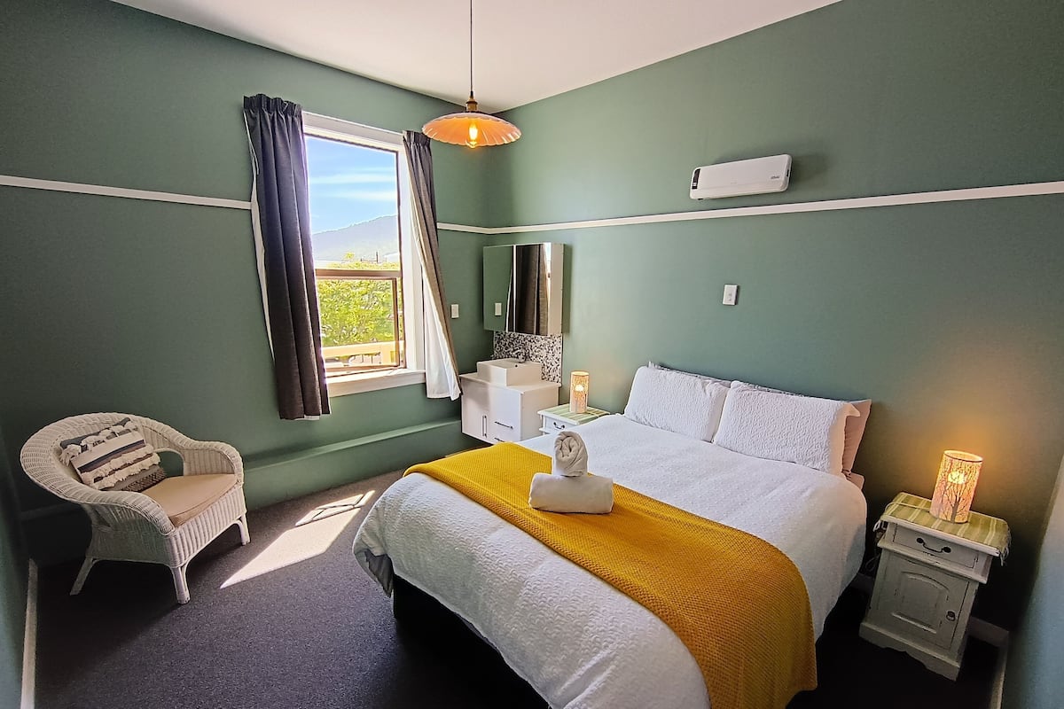 A bedroom with a bed, a chair, a bedside table with a lamp, and a window with curtains.