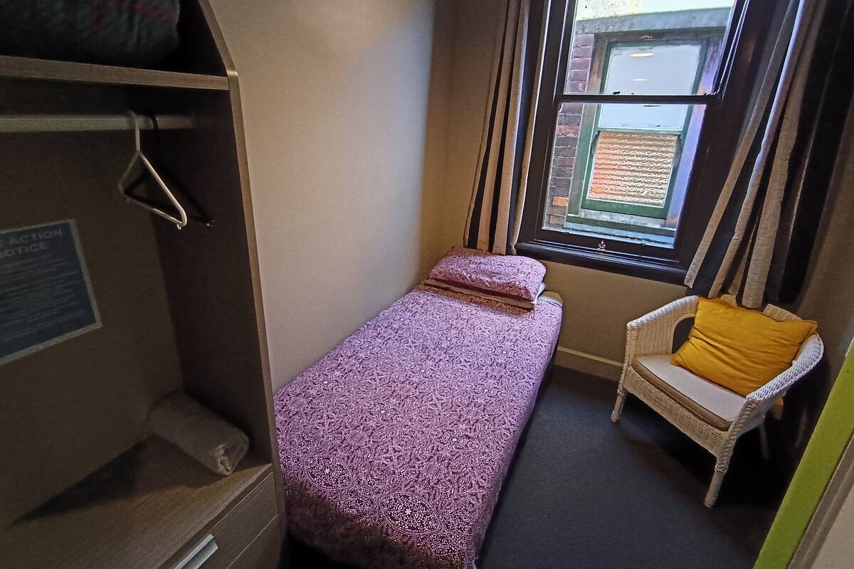 A small, compact room with a single bed, a chair with a yellow cushion, a window with curtains, and a notice on the wall.