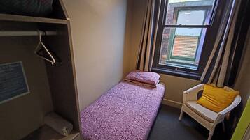 A small, compact room with a single bed, a chair with a yellow cushion, a window with curtains, and a notice on the wall.