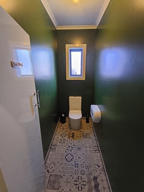 A compact bathroom with a toilet, a small window, and patterned tiles. - The Duke Hostel (Greymouth)