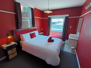 A hotel room with a bed, bedside tables, a nightstand, a window with curtains, a hanging light, a bathroom with a sink and mirror, and a view of buildings outside. - The Duke Hostel (Greymouth)
