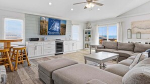 A spacious living room with a large sofa, ottomans, a dining area, and a kitchen with white cabinets. - Sandfiddler | Oceanfront | Dog Friendly, Private Pool, Hot Tub | Nags Head (Nags Head)