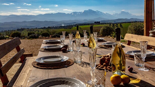 Mesa de madera lista para una comida, con botellas de vino, copas y frutas, con vistas a un paisaje montañoso.