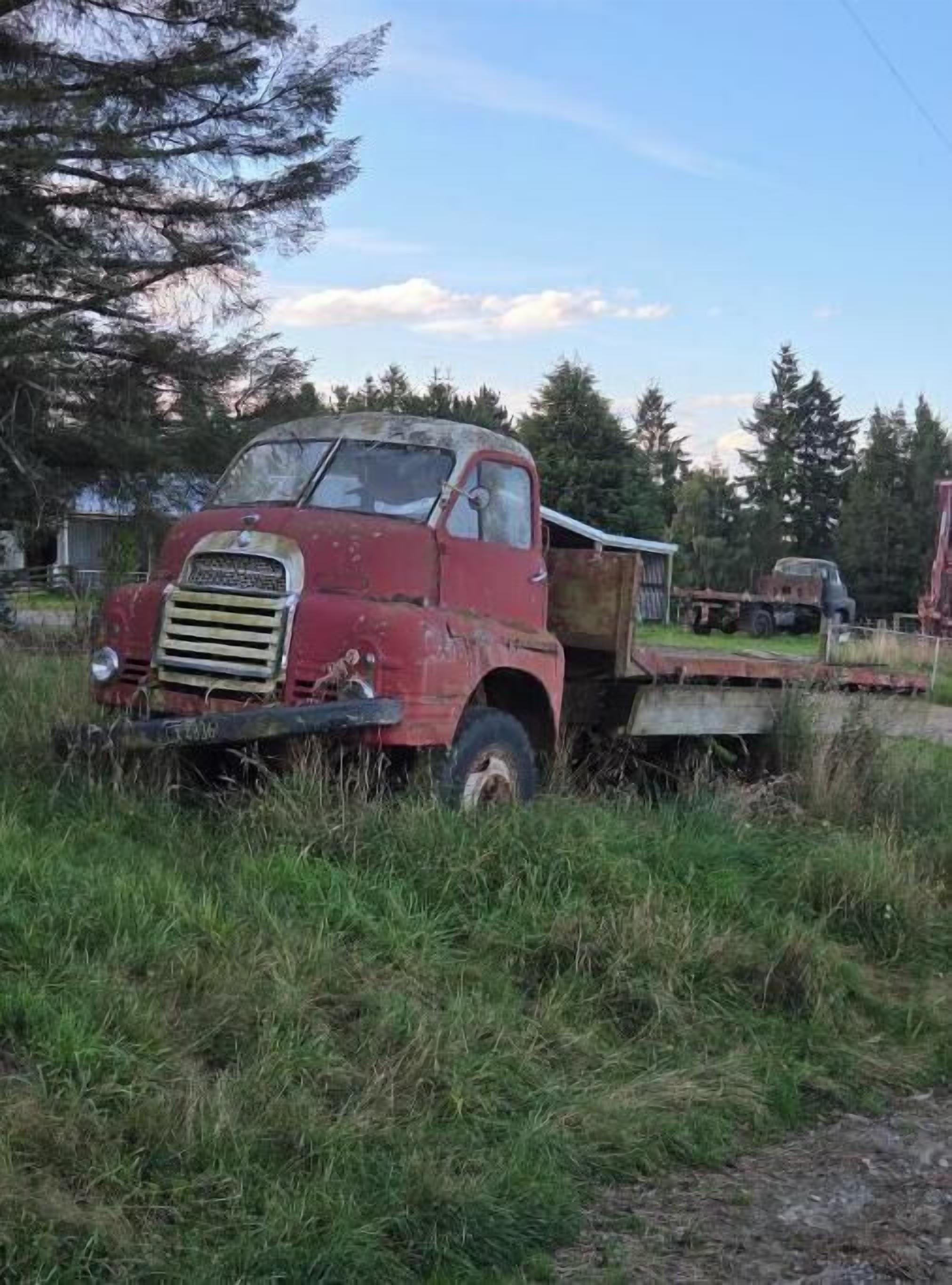 An abandoned red truck with a flatbed trailer in a grassy area.