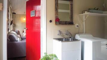 Ein kleiner Raum mit roter Tür, einem kleinen Waschbecken, einer Waschmaschine und einem Spiegel an der Wand.