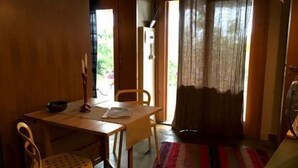 Ein Zimmer mit Holztisch, zwei Stühlen, einem kleinen Hocker, einem Bett mit gestreifter Decke, einem Fenster mit Vorhängen und einer Tür, die nach draußen führt.