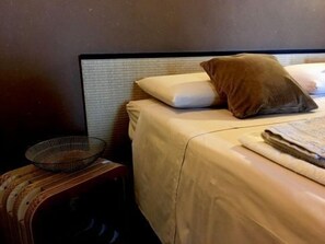 Ein ordentlich bezogenes Bett mit einem weiß-braunen Kissen, einem gefalteten Handtuch und einem kleinen Drahtkorb auf einem Holzschemel daneben.