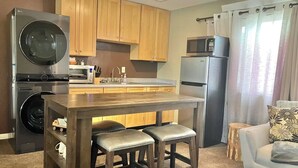 A compact kitchen with wooden cabinets, a built-in microwave, a refrigerator, and a washing machine. There is a small island with stools and a dining table. - Ravens Sunny Days King Studio w/Kitchenette, w/d, deck fire pit/grill (Westcliffe)