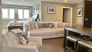 A modern living room with a sectional sofa, a dining table with bar stools, and a kitchen area in the background. - Ravens Sunny Days King Studio w/Kitchenette, w/d, deck fire pit/grill (Westcliffe)