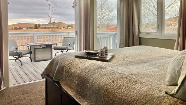 A bedroom with a large bed, a small table with two chairs, and a view of a balcony with a railing. - Ravens Sunny Days King Studio w/Kitchenette, w/d, deck fire pit/grill (Westcliffe)