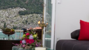 Ein Zimmer mit Blick auf eine zerklüftete Küstenlinie und Bäume. Ein Glastisch mit Kerze und Blumen ist vorhanden. Ein rotes Kissen liegt auf einem dunklen Sofa.