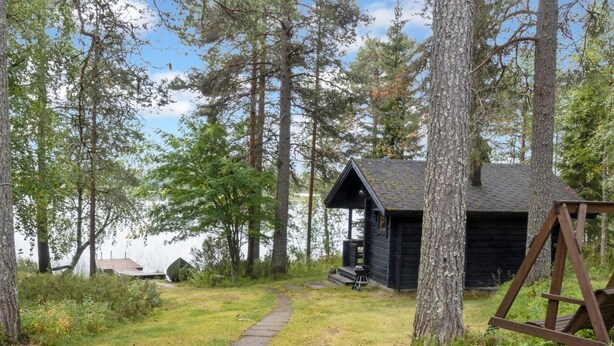 A cabin with a porch, surrounded by tall trees and a grassy area with a path leading to it.