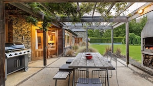 Een terras buiten met een steenbarbecue, een houten tafel met banken en een houtgestookte oven.