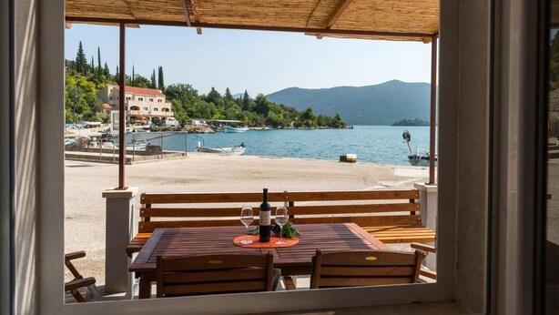 Un tavolo e una panca di legno con una bottiglia e bicchieri, affacciati su una spiaggia con barche e edifici sullo sfondo.