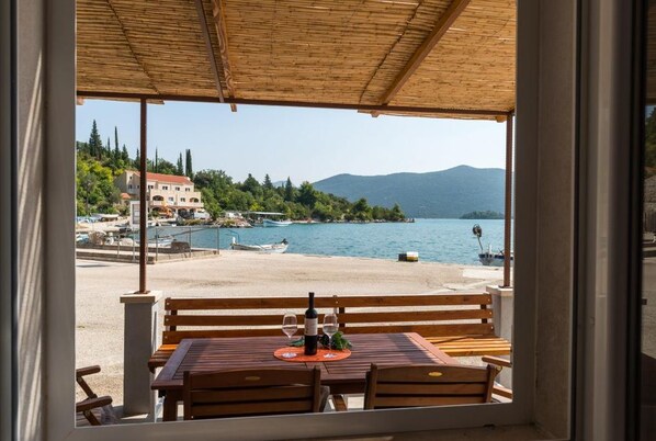 A wooden table and bench with a bottle and glasses on it, overlooking a beach with boats and buildings in the background. - Holiday Home Anima Maris- Duplex Two Bedroom Holiday Home with Terrace and Sea View (Ston)