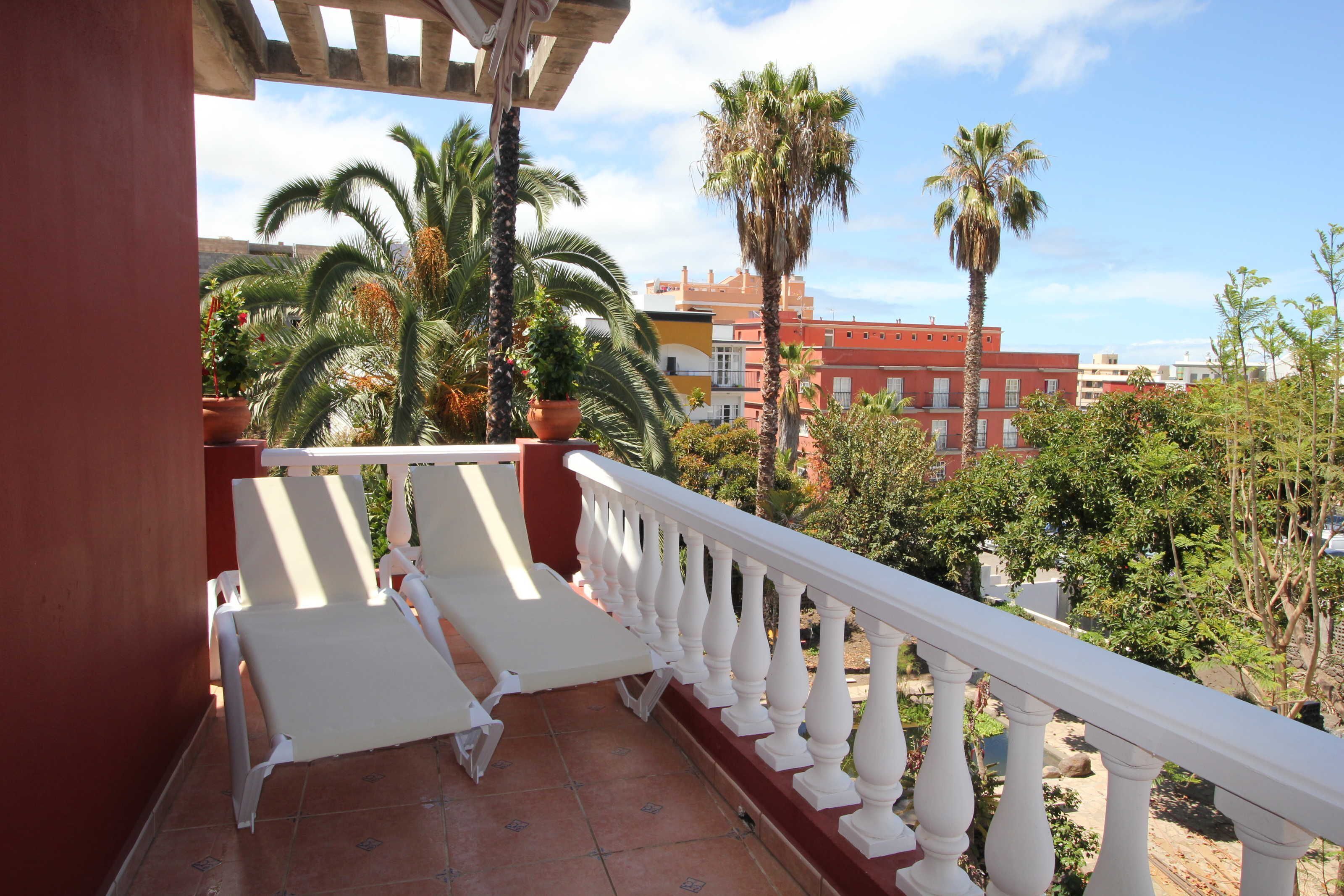 A balcony with white chairs and a white balustrade, overlooking a garden with palm trees and a red building in the background.