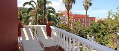 A balcony with white chairs and a white balustrade, overlooking a garden with palm trees and a red building in the background.